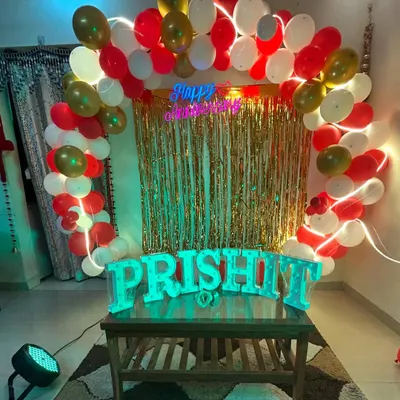 A beautiful anniversary decoration setup for a home party. It features a balloon arch in red, white, and gold, a shimmer curtain backdrop, a neon sign, and light up letters for the couple's name.