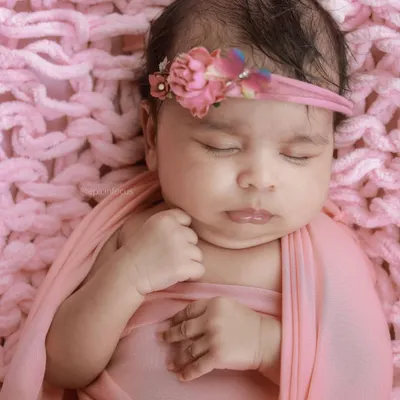 A classic newborn portrait of a baby girl in a pink swaddle and floral headband, resting on a cozy pink knit blanket.