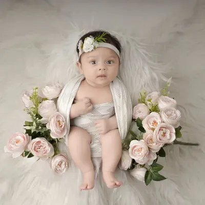 A beautiful, wide-eyed baby girl looking right at the camera. She is wrapped in white and surrounded by soft pink roses, creating a sweet and angelic portrait.