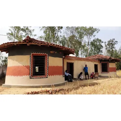 An exterior view of a community-built homestay, showcasing the rounded structure and traditional striped paintwork that reflects the local aesthetic. The building sits naturally in the landscape, a product of the earth it stands on.