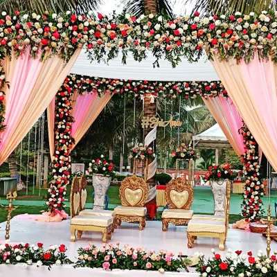 A beautiful outdoor mandap setup with pink and peach drapes, surrounded by lush greenery and palm trees.