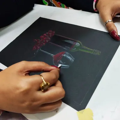 A student creating a still life of a wine bottle and glass using oil pastels on a black sheet. This technique teaches how to render light and reflection on dark surfaces.