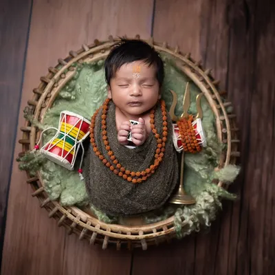 A classic 'Har Har Mahadev' shot. The baby is safely swaddled and posed in a basket, surrounded by symbolic items like the trishul and damaru, creating a serene and powerful image.