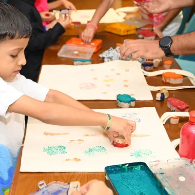 A close-up view of children's hands using colorful stamps to decorate tote bags. We use child-safe fabric paints and a variety of fun tools to make the experience tactile and exciting for young artists.