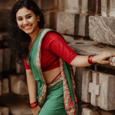 A radiant portrait of a woman in a beautiful green and red saree. Her bright, genuine smile is captured against a rustic stone wall, showcasing a natural and joyful moment.