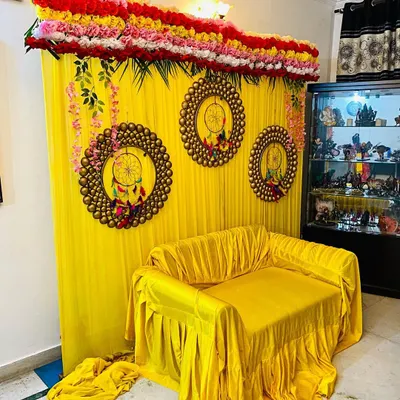 This is a popular setup for a home mehndi or haldi ceremony. It features a bright yellow fabric backdrop, a matching yellow cover for the sofa, and decorative rings to add a modern touch.