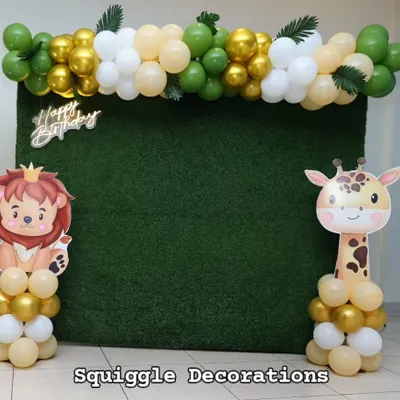 A fun jungle-themed photo booth with an artificial grass backdrop, a 'Happy Birthday' sign, and cute lion and giraffe cutouts. The green, gold, and white balloon garland adds a touch of celebration.