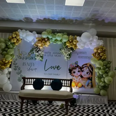 A serene and beautiful baby shower setup. I used a soft green and gold balloon arch over a backdrop featuring a lovely Radha and Krishna illustration, with the message "We're adding a little more love to our family."