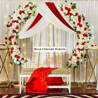 An elegant backdrop perfect for a Roka ceremony or anniversary celebration. The design features a full arch of artificial red and white roses over a white bench. The background is composed of white drapes with red accents and subtle fairy lights, creating a romantic and picture-perfect setting.