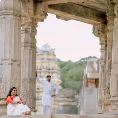 A wide shot of a couple posing amidst the stone pillars of a historic temple, with the gopuram visible in the background. This composition emphasizes the grandeur of the location.