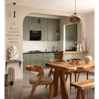 This kitchen features olive green shaker-style cabinets and a live-edge breakfast counter that brings organic charm. The entrance is framed by an elegant arch with limewash walls, and a minimal puja unit sits serenely to one side.