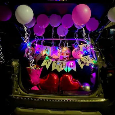 A full view of a birthday car trunk surprise. The back of the car is filled with pink and white balloons, a "Happy Birthday" banner lit by fairy lights, and two large red heart balloons.