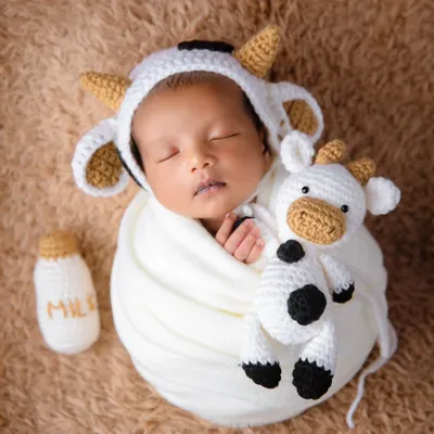 Holy cow, this is cute! A newborn dressed in a hand-knitted cow outfit, complete with a little milk bottle prop, sleeping on a soft brown blanket.
