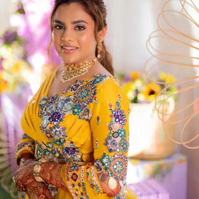 A colorful Haldi look for a vibrant bride. She is wearing a stunning yellow outfit with multi-colored embroidery, and her makeup is fresh and playful with a pop of color on the eyes.