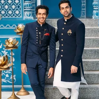 Two examples of our modern Indo-western designs. On the left, a navy blue Jodhpuri suit with a textured, darker blue waistcoat. On the right, a long navy bandhgala jacket with a gold brooch, worn over a white kurta.