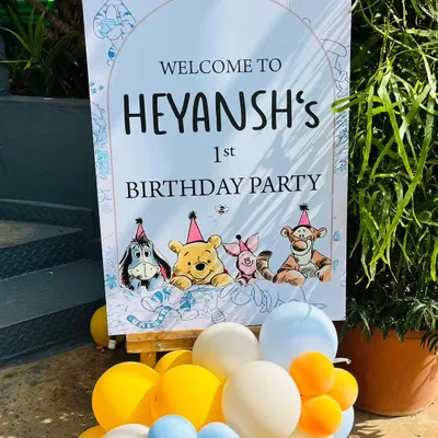A charming welcome sign for a Winnie the Pooh first birthday party. The sign features characters from the Hundred Acre Wood and is displayed on an easel with a small balloon cluster.