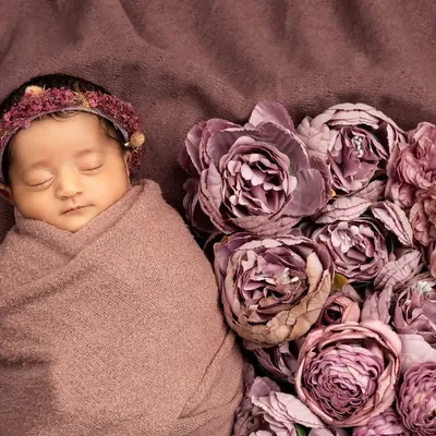 A classic newborn portrait. The baby is safely swaddled and sleeping peacefully next to a heart-shaped arrangement of purple flowers.