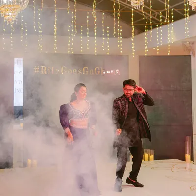 The couple's first dance at their Sangeet, surrounded by theatrical smoke. The decor features elegant chandeliers and fairy lights for a glamorous feel.