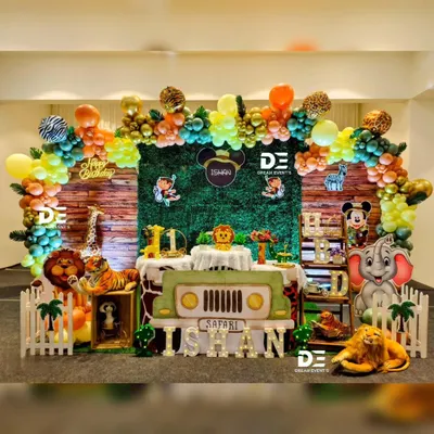 A full safari adventure stage for Ishan's first birthday. The setup includes a grass-green backdrop, a jeep-shaped table, and cutouts of all his favorite jungle animals like the lion, elephant, and monkey, all under a colorful balloon arch.