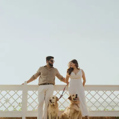 A beautiful family portrait of a couple with their two golden retrievers, posing by a white fence against a clear sky in Goa. This shot is a perfect example of a relaxed, pet-friendly pre-wedding shoot.