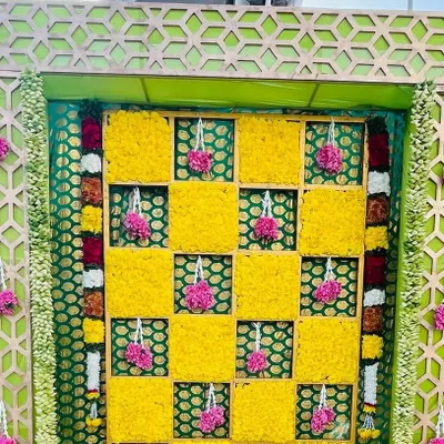A close-up of a Haldi backdrop, showing the checkerboard pattern of yellow marigolds and hanging pink floral tassels. The intricate design adds a beautiful texture to the decor.