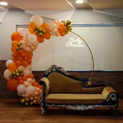 We create a special spot for the mom-to-be with this modern baby shower setup. It features a lush organic balloon arch in orange, white, and gold, a custom "Baby Shower" neon sign, and an elegant chaise lounge for comfort and perfect photos.