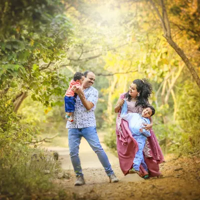 A family portrait alive with motion and joy. The father lifts the baby, the mother holds her older son, and the sunlight filtering through the trees adds a touch of magic to this candid moment.