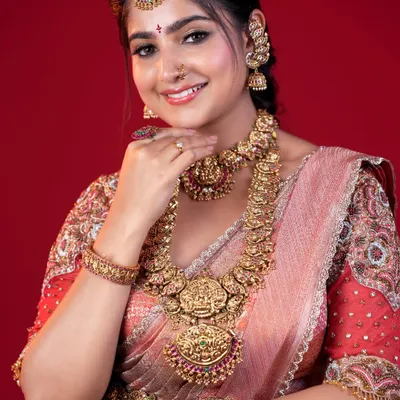 A classic South Indian bridal portrait. This look features a flawless matte base, beautifully defined eyes, and traditional temple jewelry against a vibrant red backdrop, capturing the pure essence of a muhoortham bride.