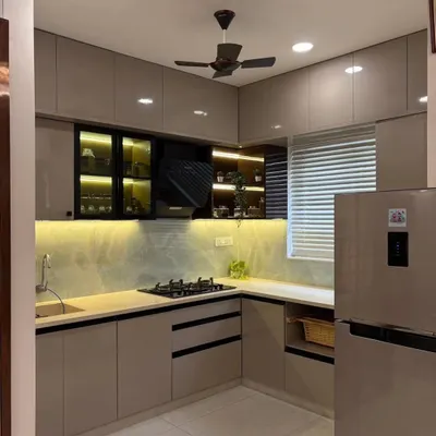 This L-shaped modular kitchen is designed for efficiency and style. It features handleless cabinets in a modern beige tone, a built-in hob, and under-cabinet lighting that illuminates the workspace. The black-framed glass cabinets add a touch of contrast and provide display storage.