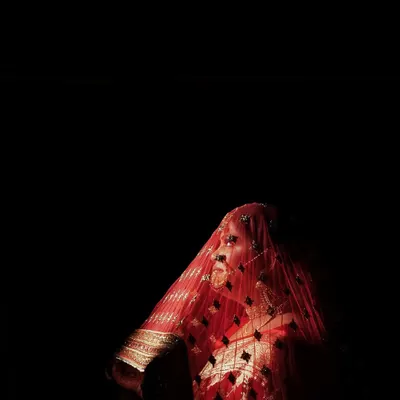 We use dramatic lighting to create powerful and artistic bridal portraits. This low-key photograph highlights the bride's silhouette and the rich texture of her red veil against a dark background, creating a truly memorable image.