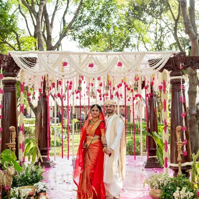 A beautiful couple framed by the grandeur of their wedding mandap. The decor is a blend of tradition and elegance, with rich wooden pillars, a ceiling of white floral strings, and accents of pink, creating a perfect backdrop for their special day.