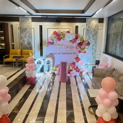 A wide shot of a 20th birthday decoration in a party hall, featuring a "Goodbye Teens, Hello Twenties" theme with pink, red, and gold balloons.