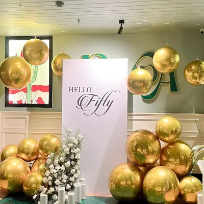 For this elegant 50th birthday, we created a stunning photo corner. The setup features a clean white backdrop with "Hello Fifty" in script, surrounded by reflective golden orb balloons of various sizes for a chic, celebratory look.