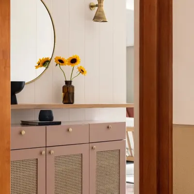 The foyer features a custom-designed console in a dusty rose color with cane panels. A large circular mirror and brass wall sconce complete this welcoming entryway vignette.