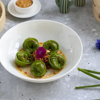 This is an example of modern, artistic food plating. The vibrant green of the spinach dumplings pops against the clean white bowl, while the careful placement of garnishes highlights the dish's elegance for a fine-dining menu.