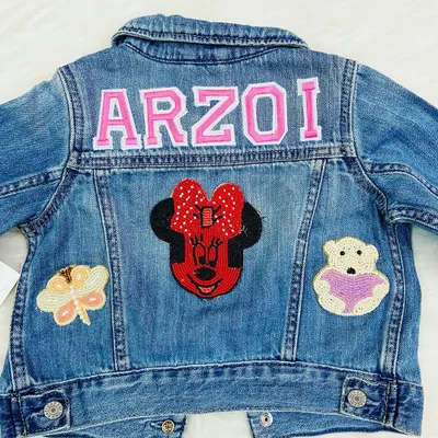 A personalized denim jacket for a little girl named 'Arzoi'. The jacket is decorated with her name in pink letters, a large Minnie Mouse patch, and other cute beaded patches.