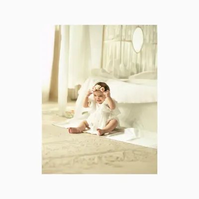 A happy baby girl sits on the floor in a bright white studio, playing with her floral headband. The clean, minimalist setting puts all the focus on her adorable expression and joyful spirit.