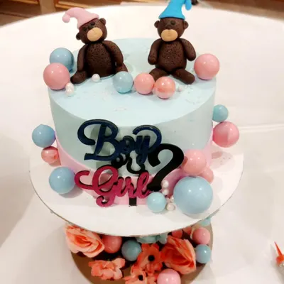 A beautiful two-tier cake for a "Boy or Girl?" gender reveal party. It features fondant teddy bears in pink and blue hats, surrounded by matching decorative balls.