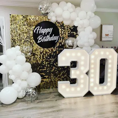 A chic and modern 30th birthday setup. The combination of a gold sequin wall, all-white balloon decor, disco balls, and bold marquee numbers creates a high-contrast, glamorous look perfect for a memorable party.