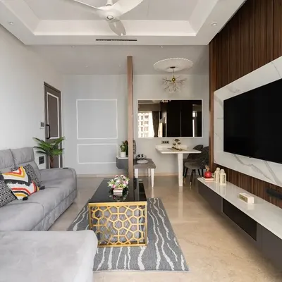 This living room combines a comfortable grey sectional sofa with a stunning TV unit featuring a marble-finish backdrop and wood fluting. The design is minimalist yet full of texture and warmth, perfect for a modern family. This is the wide shot of the space.
