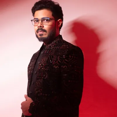 A stylish portrait of the groom-to-be against a red background. His direct gaze and sharp attire convey a sense of modern confidence.