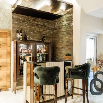 A luxurious home bar setup with Travertine marble walls, a dark wood ceiling, and plush forest-green velvet bar stools.