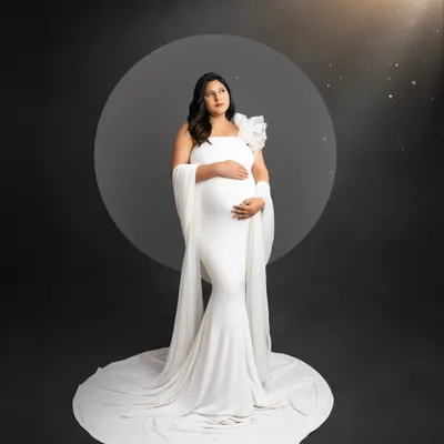 A classic and elegant solo maternity portrait. The mom-to-be wears a simple white gown, standing on a white circular platform against a dark background, with a soft light creating a celestial glow.