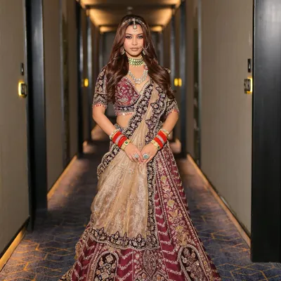 A full-length portrait of the modern maharani look, showcasing the deep maroon lehenga with intricate golden embroidery. The makeup is balanced to be impactful without overpowering the grandeur of the traditional attire.
