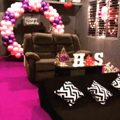 A popular birthday setup at one of our Bengaluru locations, featuring a purple and pink balloon arch and custom initial lights. The room has a mix of recliner and floor seating with comfy cushions.