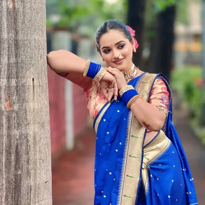 A beautiful outdoor shot of a model in a traditional blue Paithani saree. The makeup is kept classic with a prominent nath and choker, perfect for a pre-wedding photoshoot.