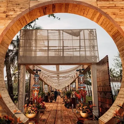 The dramatic circular wooden entrance to the 'Bora Bora' themed event, perfectly framing the long, inviting pathway lined with Tiki statues and fairy lights. This sets the stage for a journey into a tropical wonderland.
