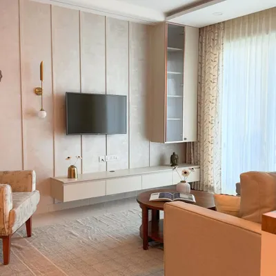 A living room that whispers elegance. The design uses soft beige tones, a floating TV unit with metallic accents, and warm lighting to create a serene, light-filled retreat.