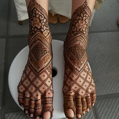 A stunning example of full-coverage feet mehendi. This design combines intricate floral bands around the ankle with a bold geometric diamond pattern that covers the entire foot.