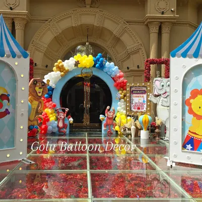 The entrance to a carnival-themed birthday party, featuring a colorful balloon arch and circus animal cutouts over a glass floor filled with flower petals.